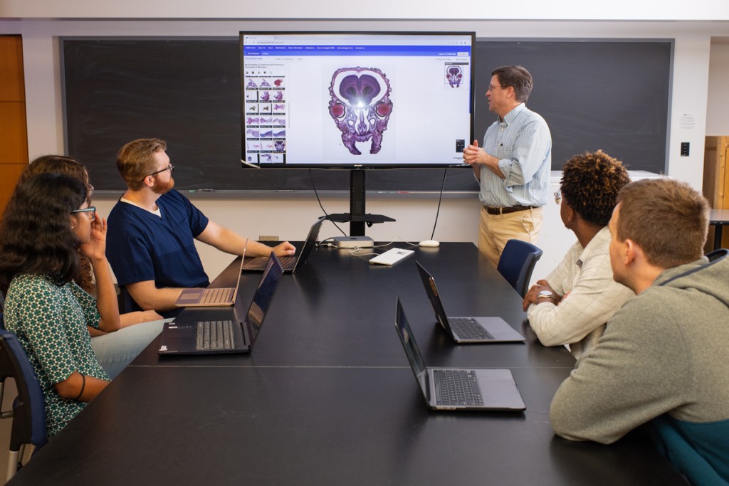 An anatomy professor standing in front of a classroom with four students stands in front of a screen with anatomical images