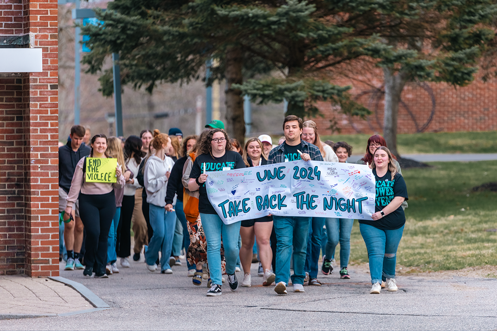 A group of students marches on campus holding a sign that reads "UNE 2024 Take Back the Night"