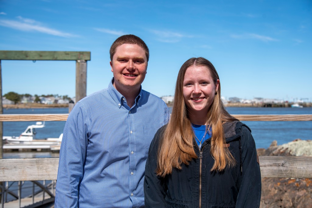 Two people pose on a dock and smile in front of the ocean