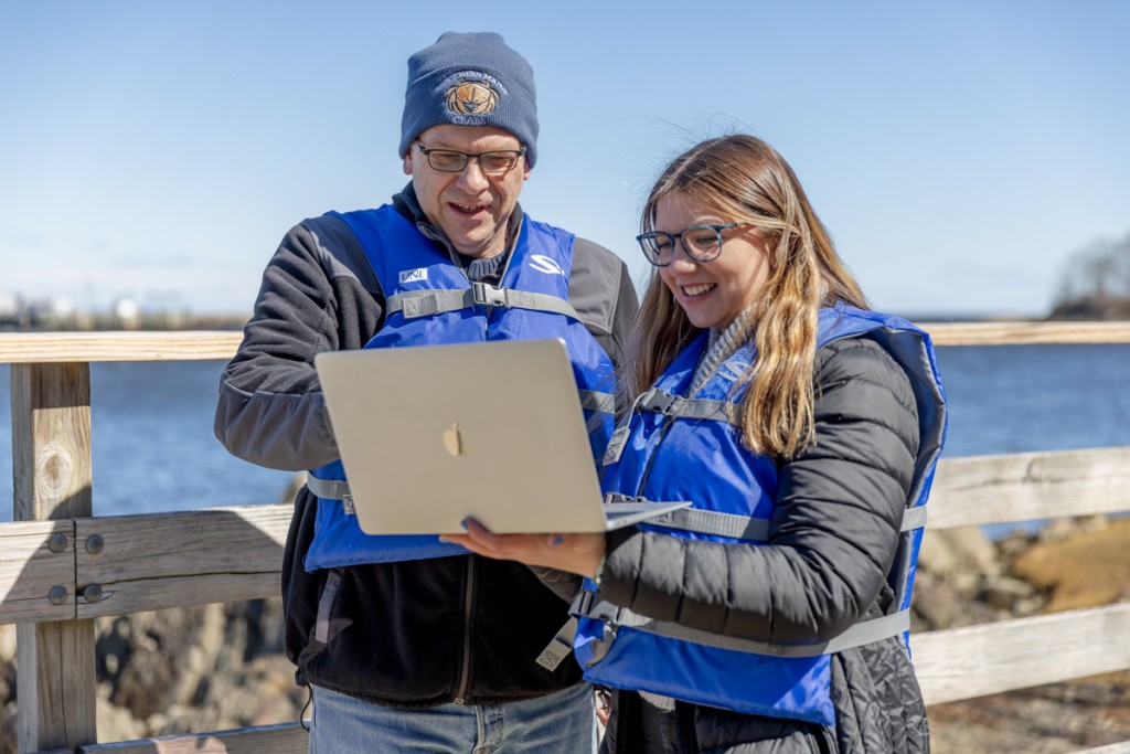 A student reviews data on a laptop with Professor Markus Frederich while both wear life jackets and stand by the ocean