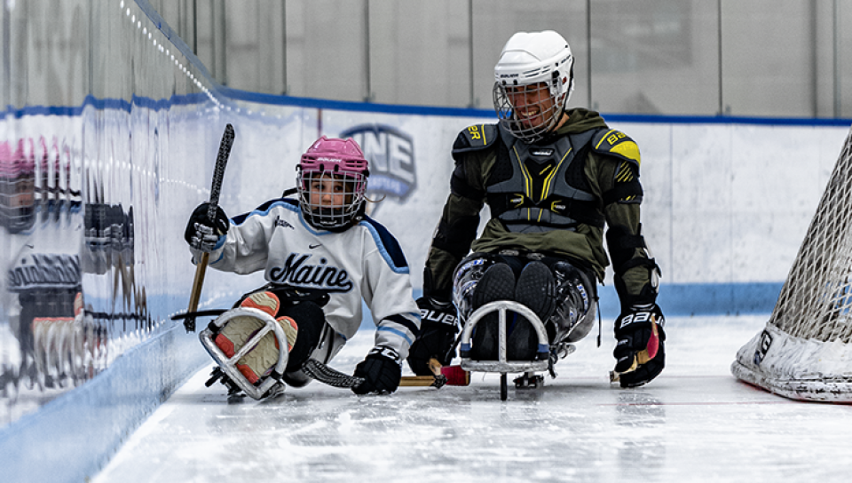 A man and child round the corner using adaptive sleds on the UNE ice hockey rink