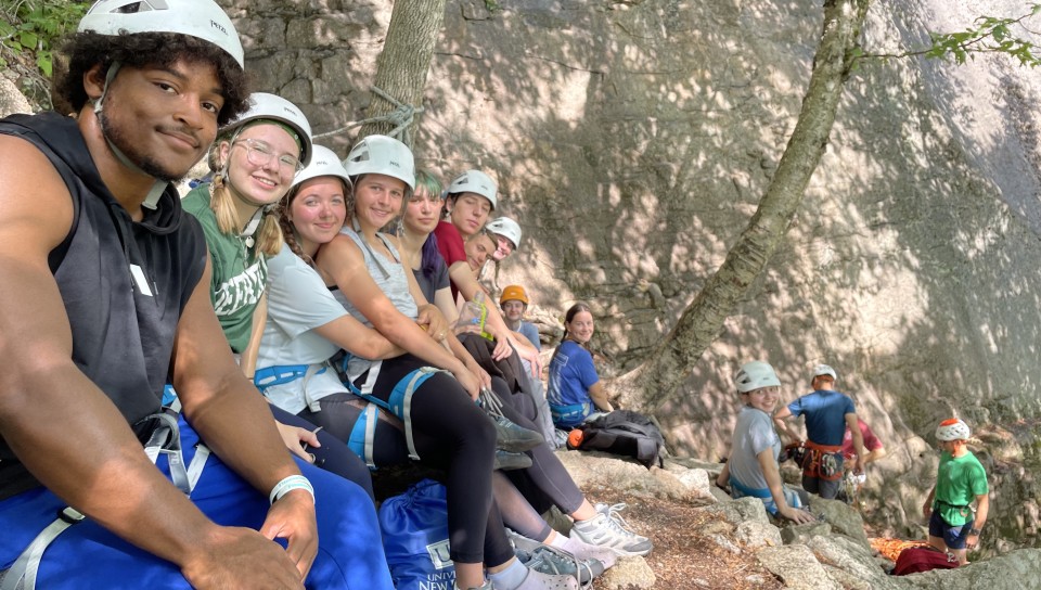 Several students, wearing helmets, take a selfie over the side of a mountain