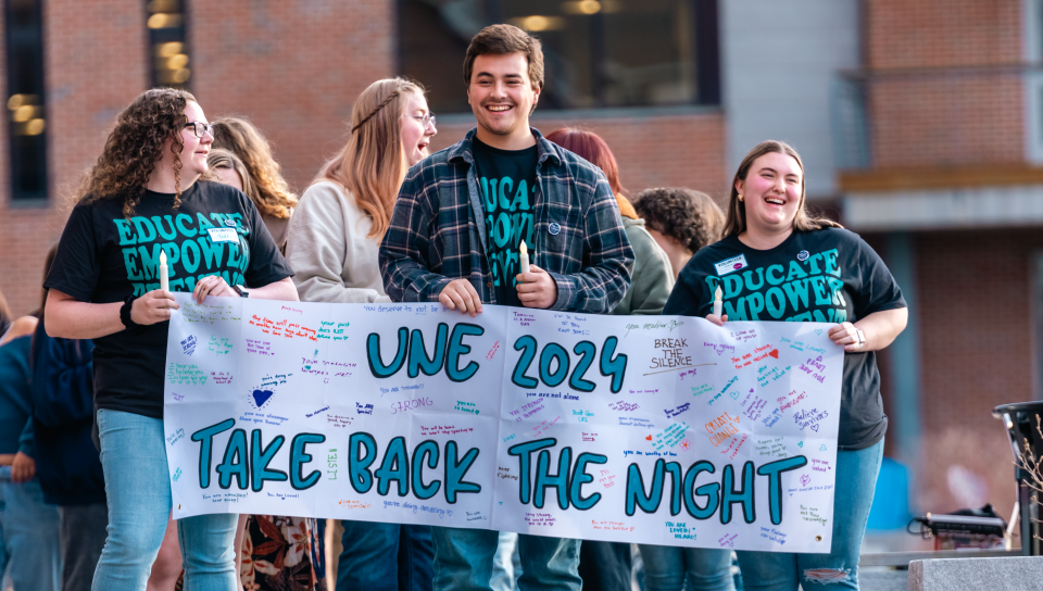 Three students hold up a banner that reads "UNE 2024 Take Back the Night"