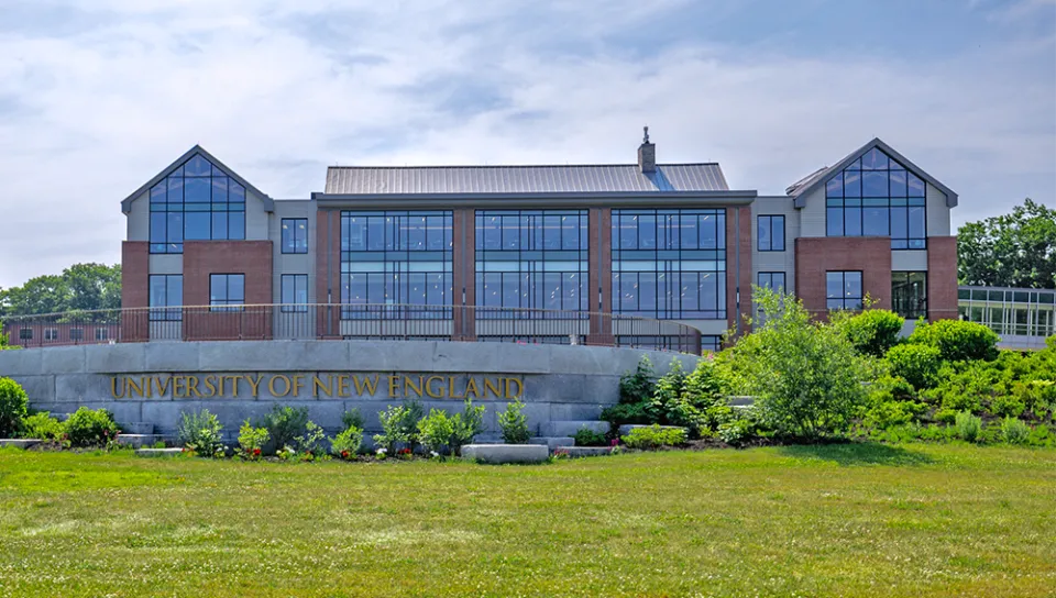 A brick building with numerous windows sits behind a stone sign that reads, "University of New England"