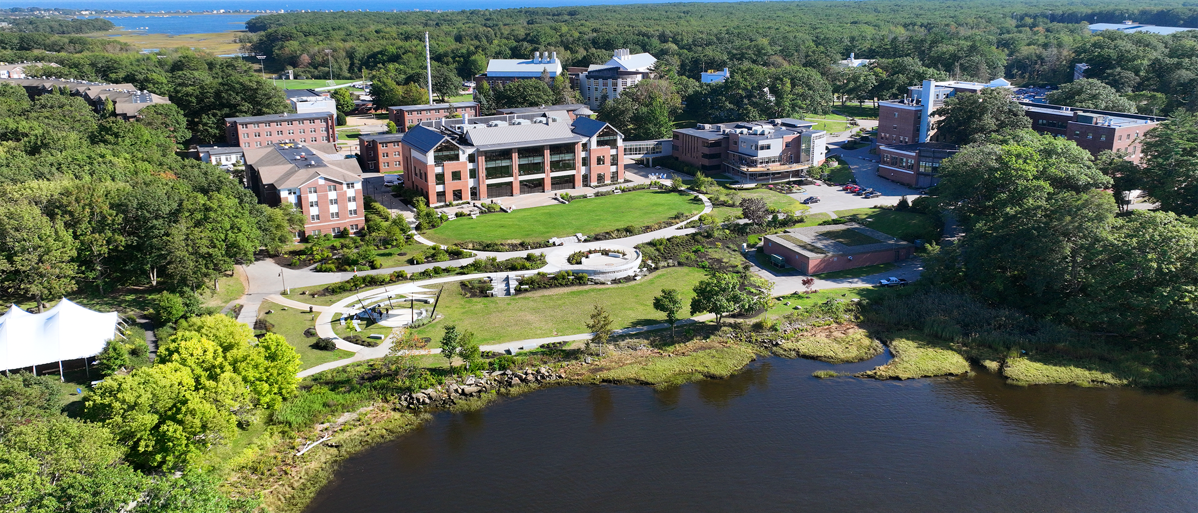 An aerial view of brick buildings with numerous glass windows sitting on green grass in front of a river