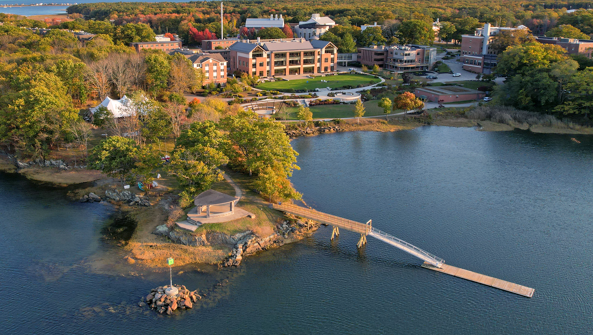 Aerial view of UNE's Biddeford campus in autumn, showing the waterfront campus with brick academic buildings, a small island with pavilion connected by a wooden footbridge, surrounded by colorful fall foliage and the Saco River estuary.