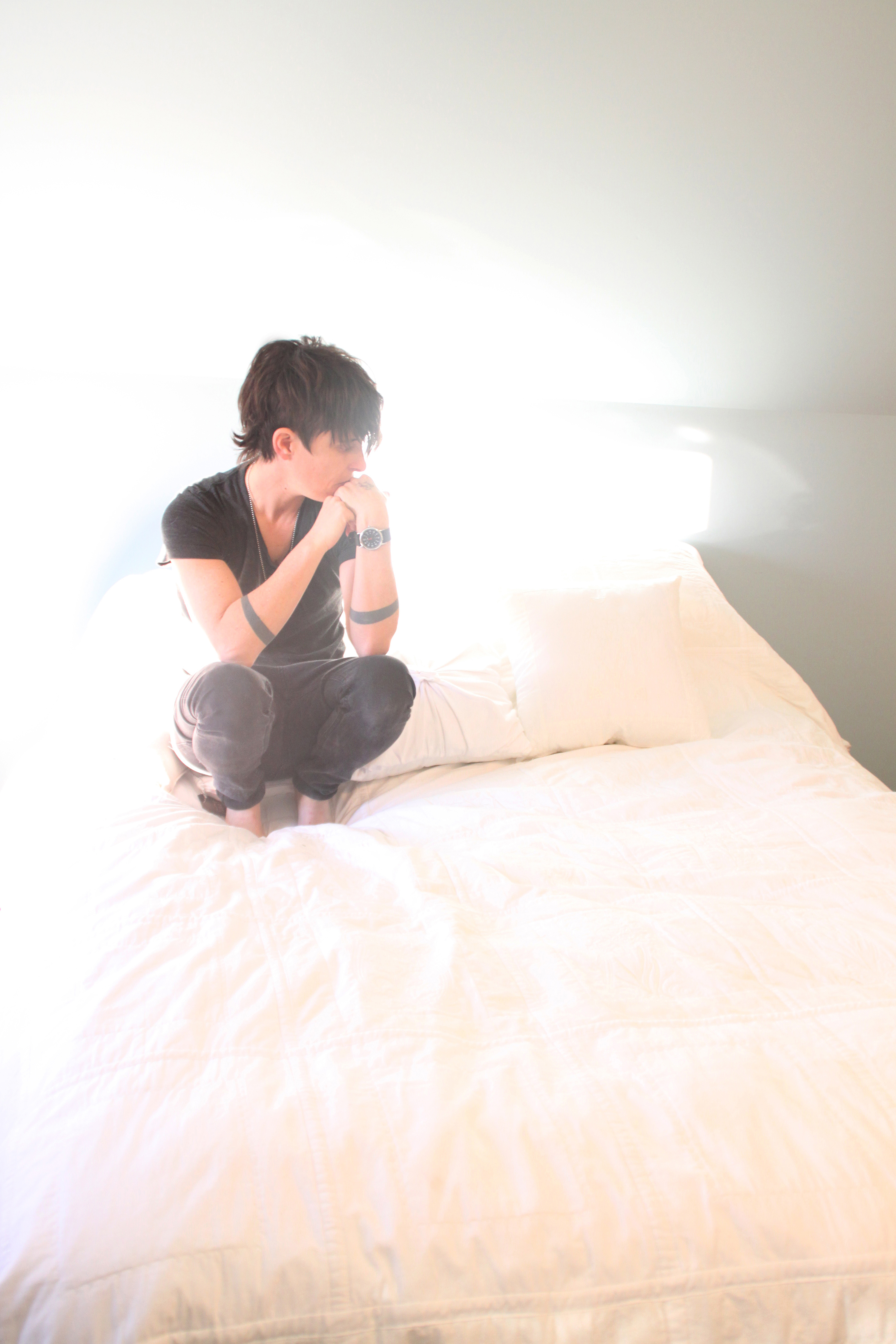 Artist Andrea Gibson crouched on bed in brightly-lit room looking away
