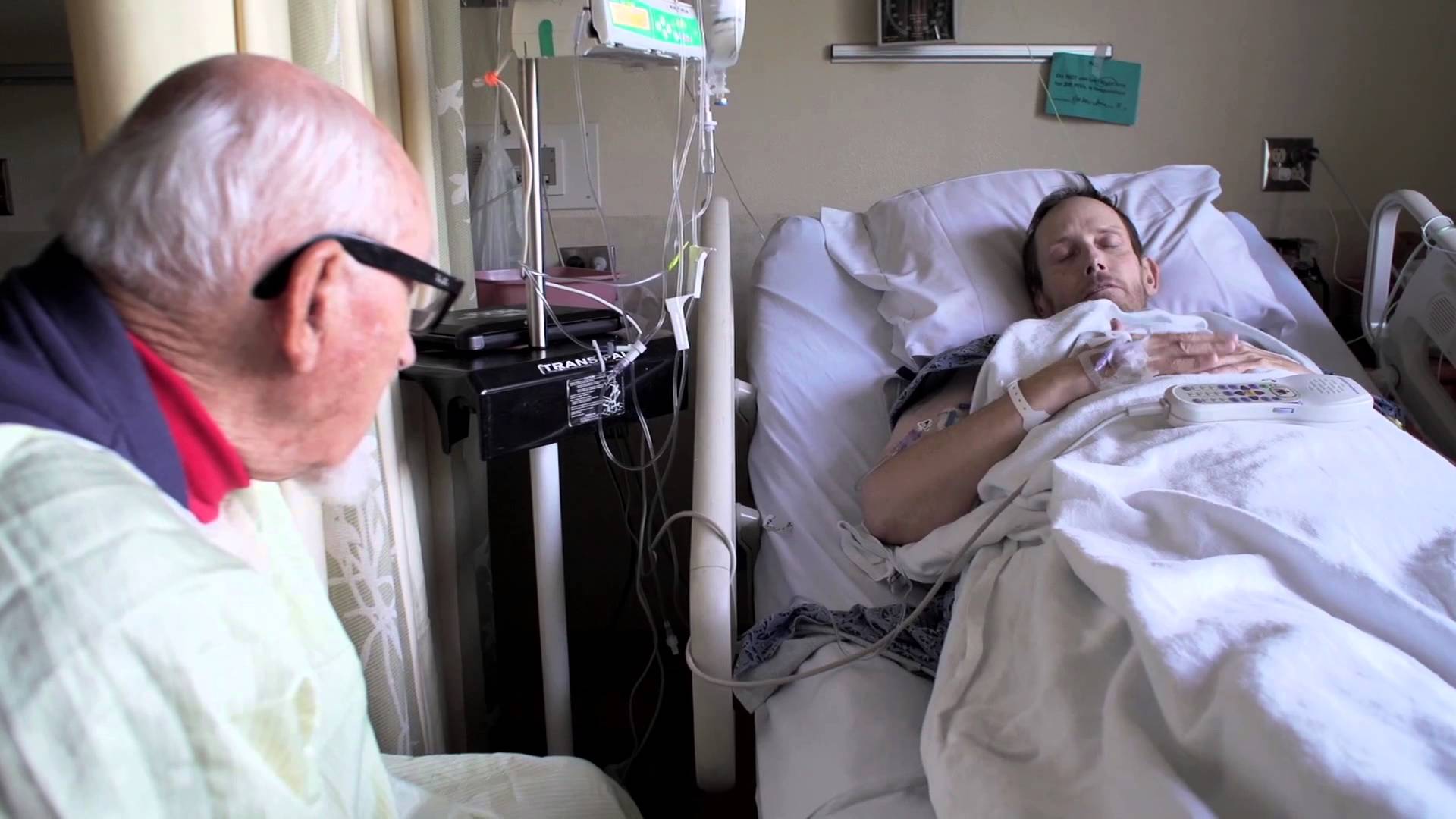 An elderly white doctor looks at a young white man in a hospital bed. 