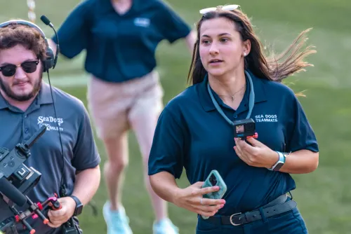 A sports management student wearing a Sea Dogs polo shirt runs next to a cameraman on Hadlock Field