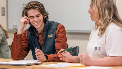 Two students talk while sitting at a desk with paperwork and notebooks in front of them