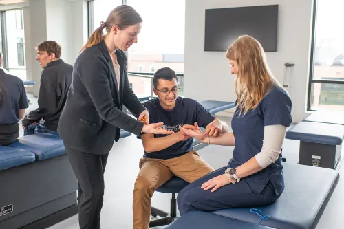 An O T professor assists a student as he uses a rubber band to learn about muscle activation and movement on another student's wrist