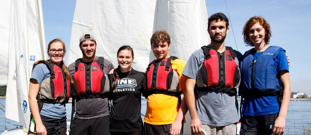 a group of students wearing lifejackets pose in front of sailboats in the water