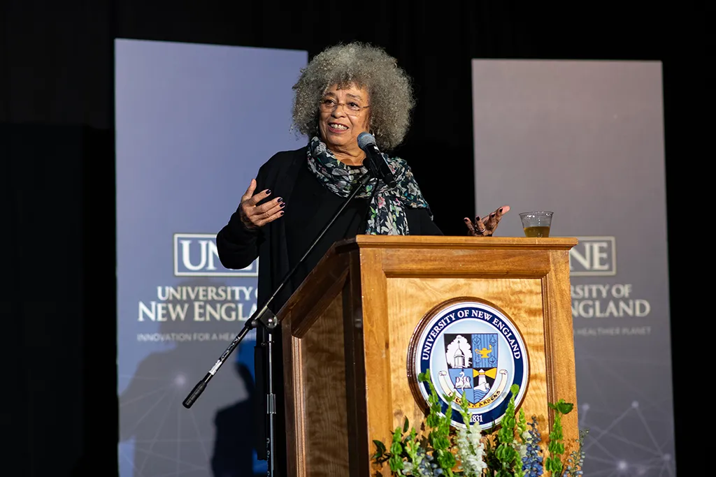 Angela Davis stands at a podium in front of a screen displaying the U N E logo