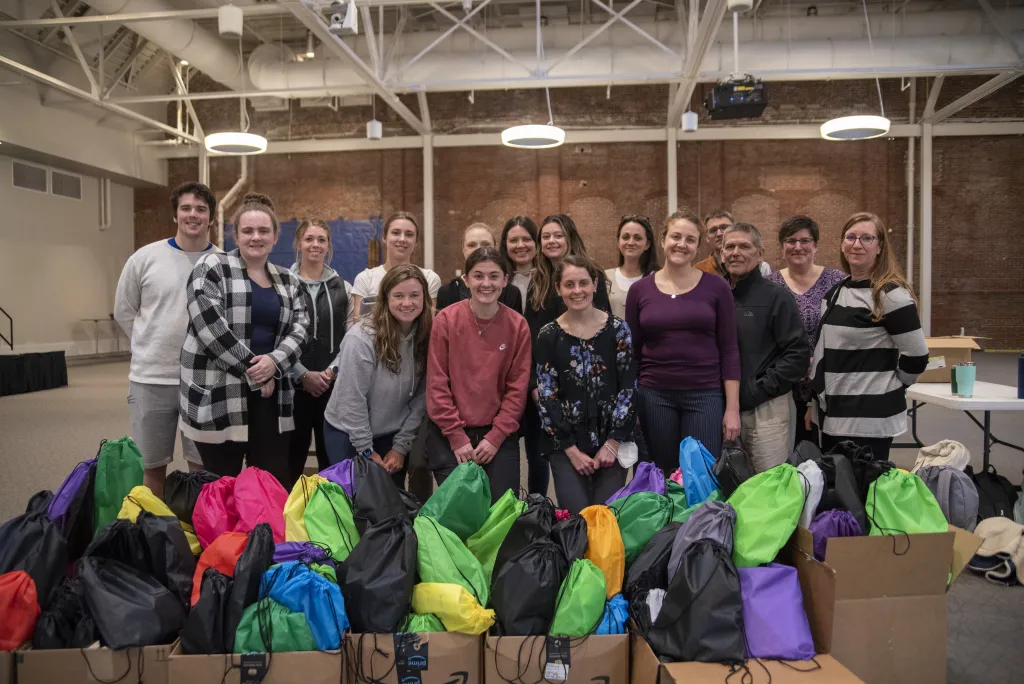 12 students and guests stand in front of harm reduction bags assembled for released inmates