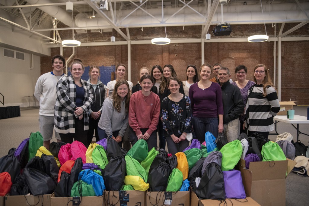 12 students and guests stand in front of harm reduction bags assembled for released inmates
