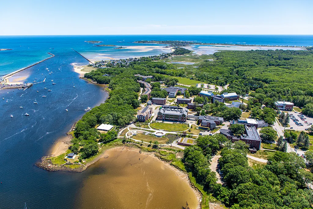 An aerial view of the Biddeford Campus showing campus buildings, nearby forest, beach, river, and the ocean