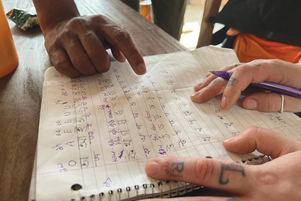 A close-up of two individual's hands pointing to parts of the written Bangla language in a notebook