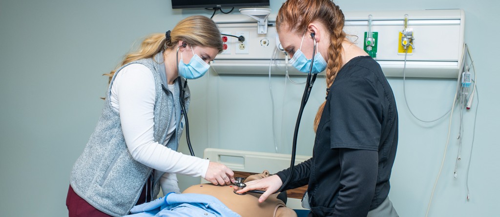 Two nursing students practicing listening to the heart and lungs with a stethoscope on a dummy in the Simulation Lab
