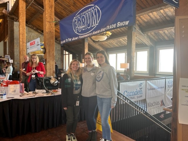 Three woman students pose for a photo in front of a banner at the Maine Fisherman's FOrum