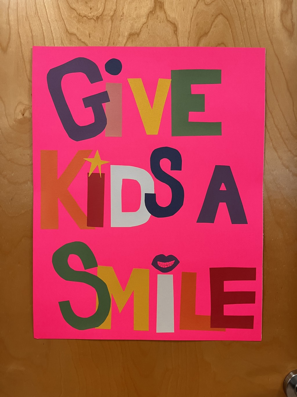 A pink poster against a wooden door reads "Give Kids a Smile"