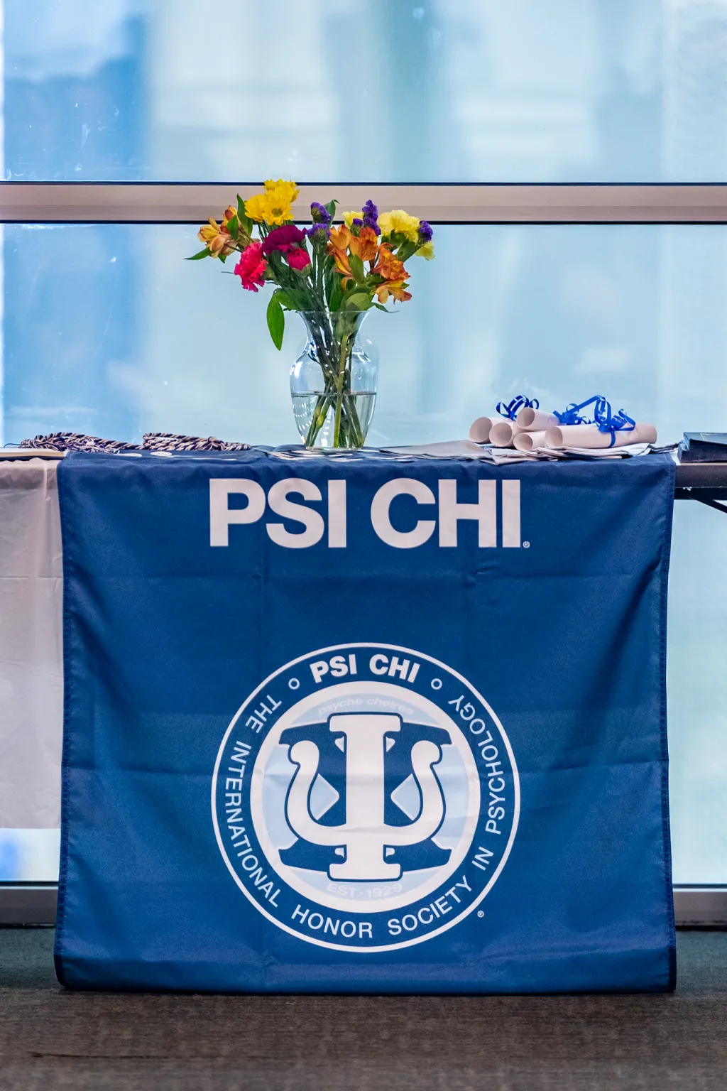 Flowers sit in a vase on a table adorned with the PSI CHI logo