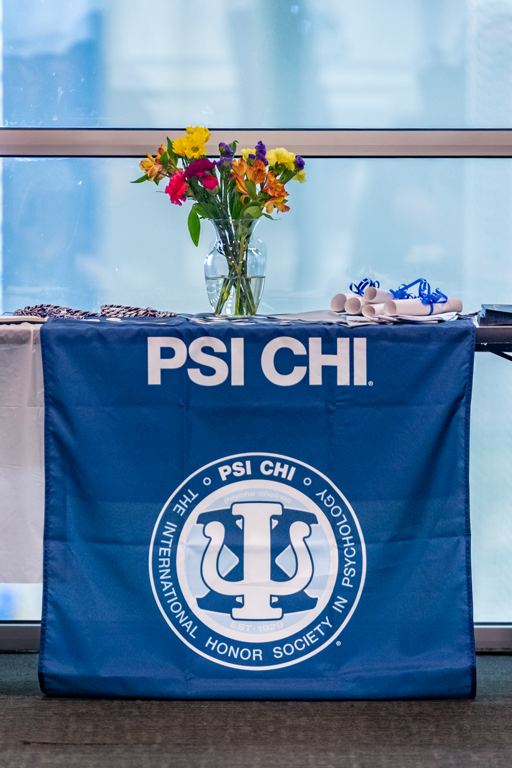 Flowers sit in a vase on a table adorned with the PSI CHI logo 