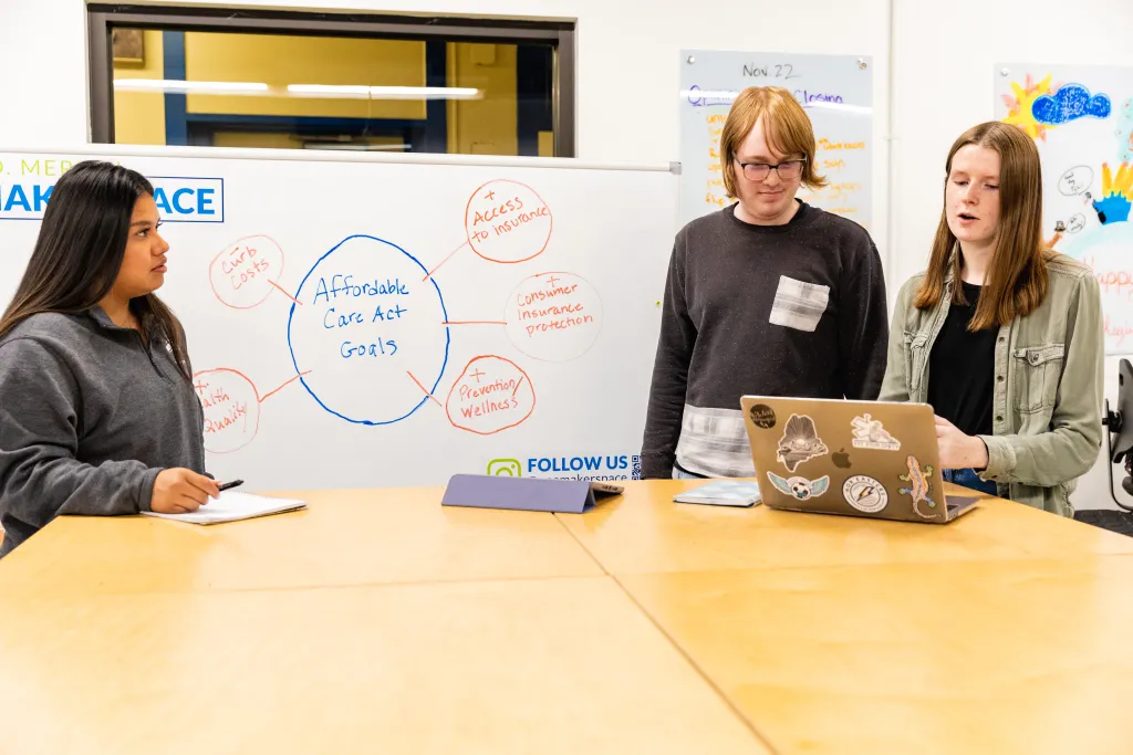 Three U N E students stand at a table in front of a whiteboard
