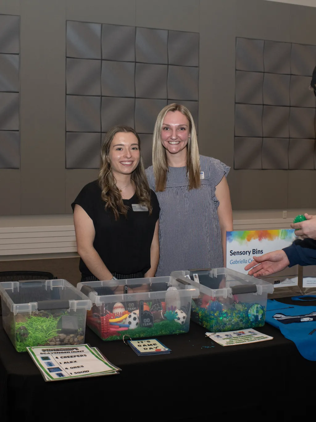 Two female students pose in front of their project, sensory bins