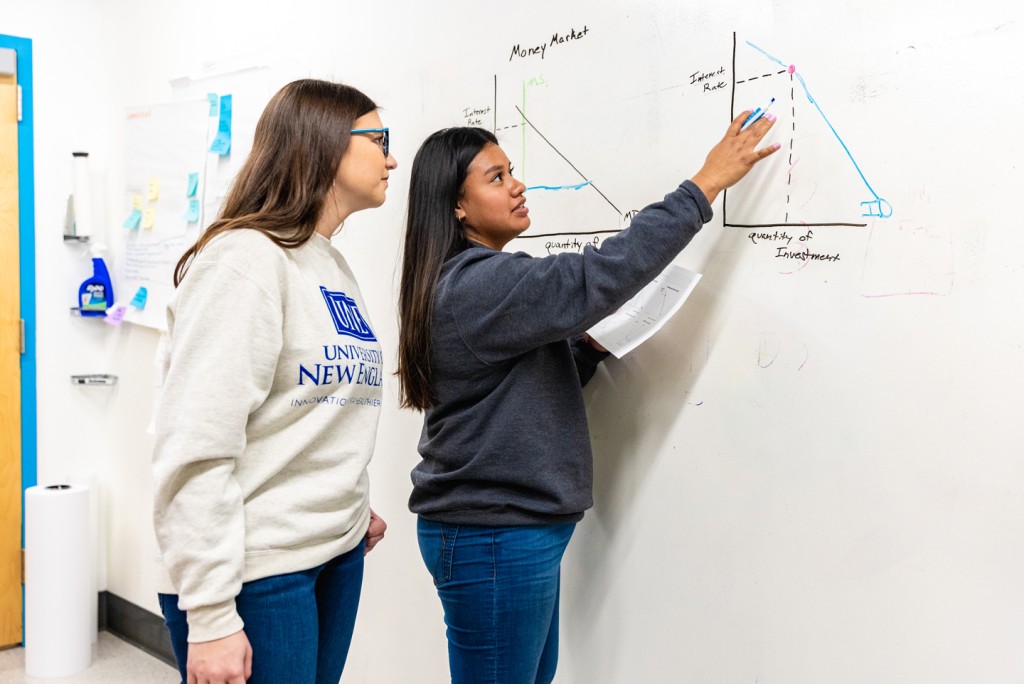 A U N E student explains a graph drawn on a whiteboard, titled "Money Market," to a fellow student