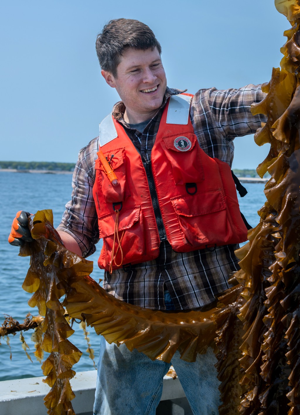 A student in a life vest stands on a boat in the ocean and holds a large piece of kelp