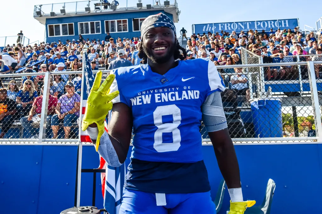 A football player smiles at the camera while standing in front of a crowd at a game