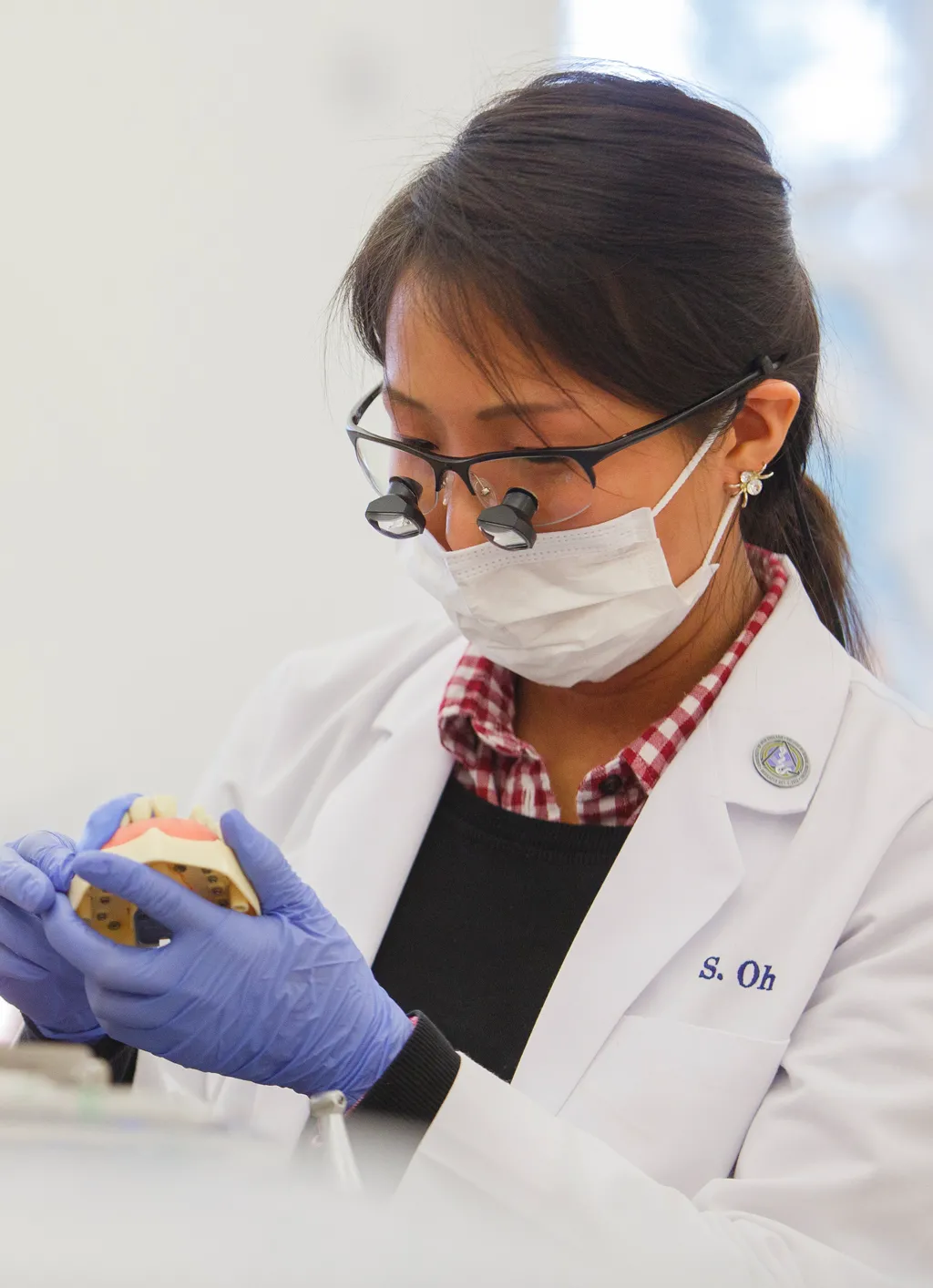 A student wearing a white coat, gloves, mask, and loupes inspects a model of teeth