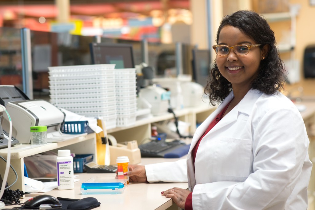 A pharmacy student in a white coat and smiling at the camera stands behind a counter in a pharmacy