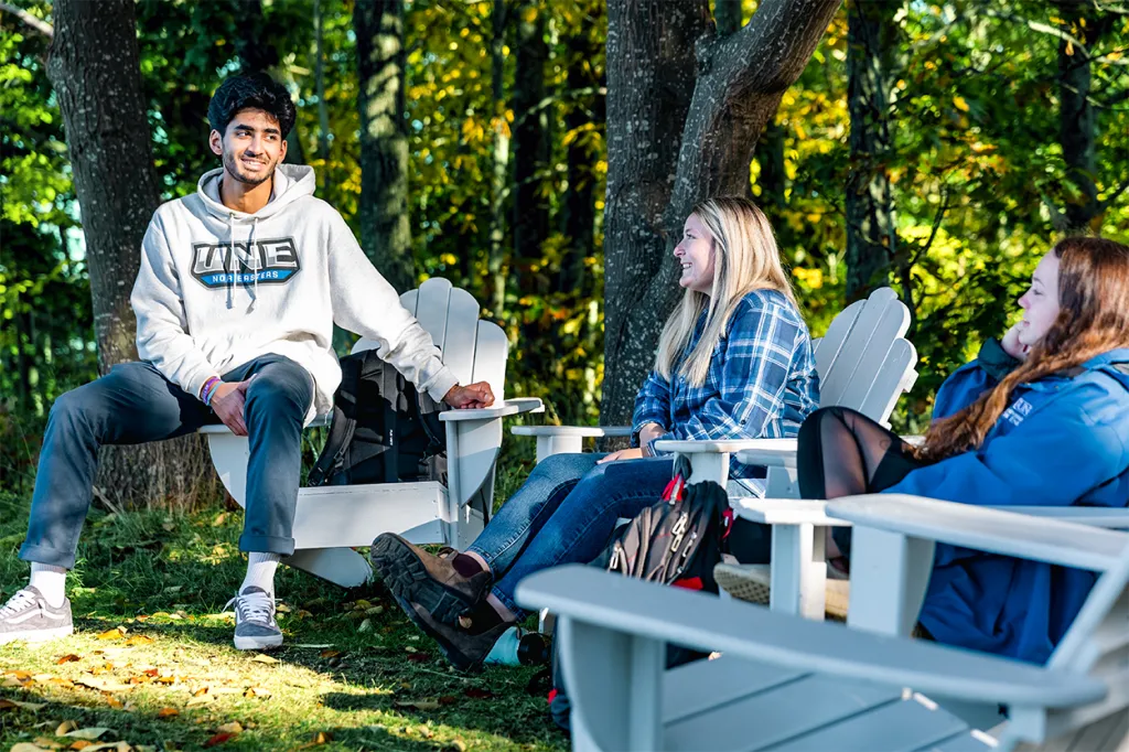 Three students sitting in adirondack chairs outside in front of the woods
