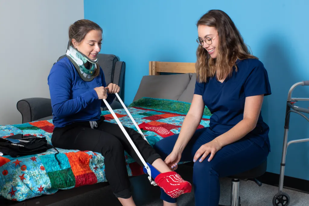 An Occupational Therapy student wearing blue scrubs assists a patient wearing a neck brace with a foot sling