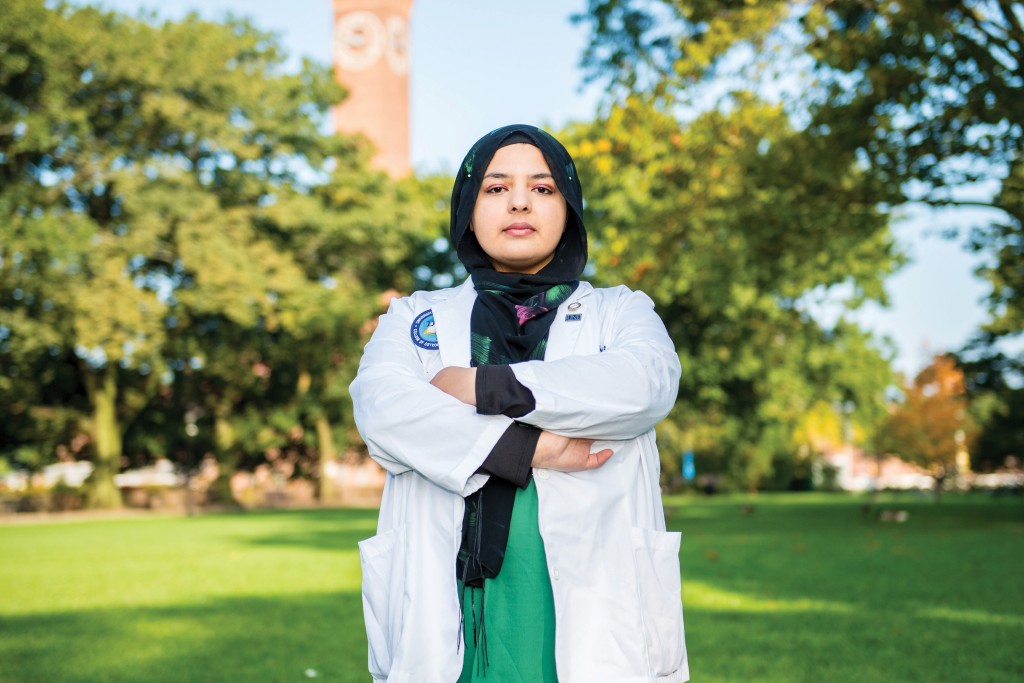 Fajar in her hijab and U N E C O M white coat with arms crossed, standing outside on a bright summer day
