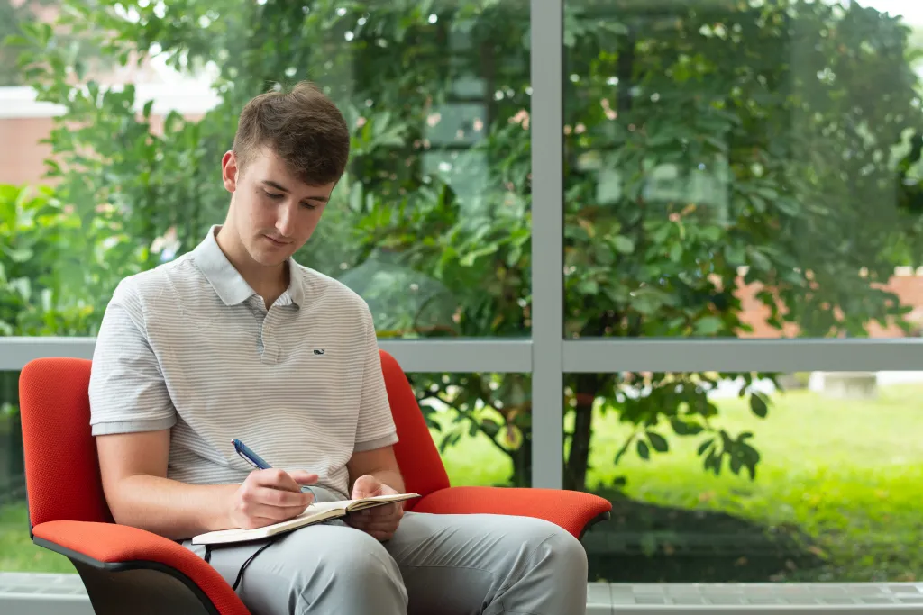U N E student Dawson Durcotte writes a poem sitting in a chair in the Biddeford campus library