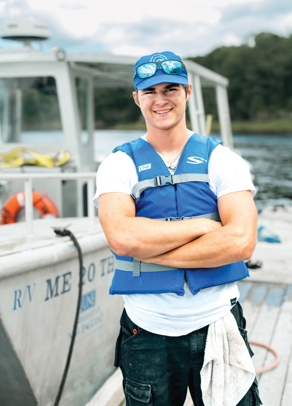 Clayton wears a blue life fest and matching baseball hat as he stands with his arms crossed in front of a U N E boat