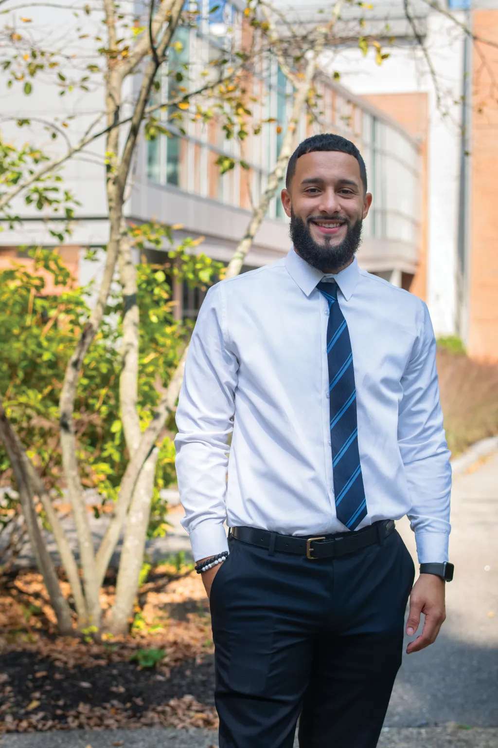 Jeremiah wearing a button down and tie as he stands in front of the glass and brick of the Commons building