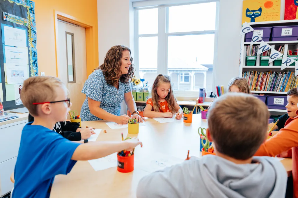 Emily and several children drawing on paper with crayons at a large classroom table