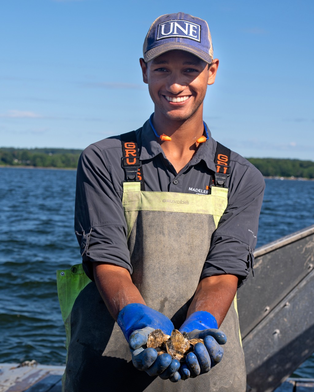 A student wearing a U N E baseball hat and waders shows off several oysters in their hands
