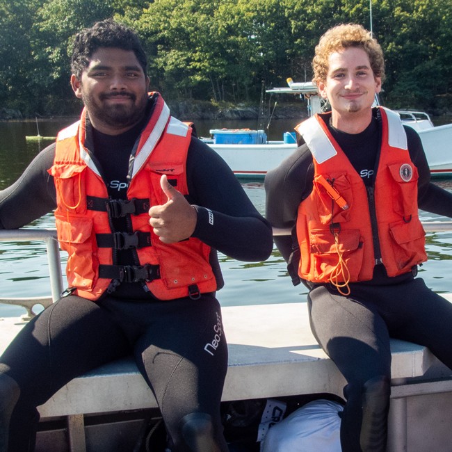 Two students in life vests smile as they sit on a bench of a U N E boat
