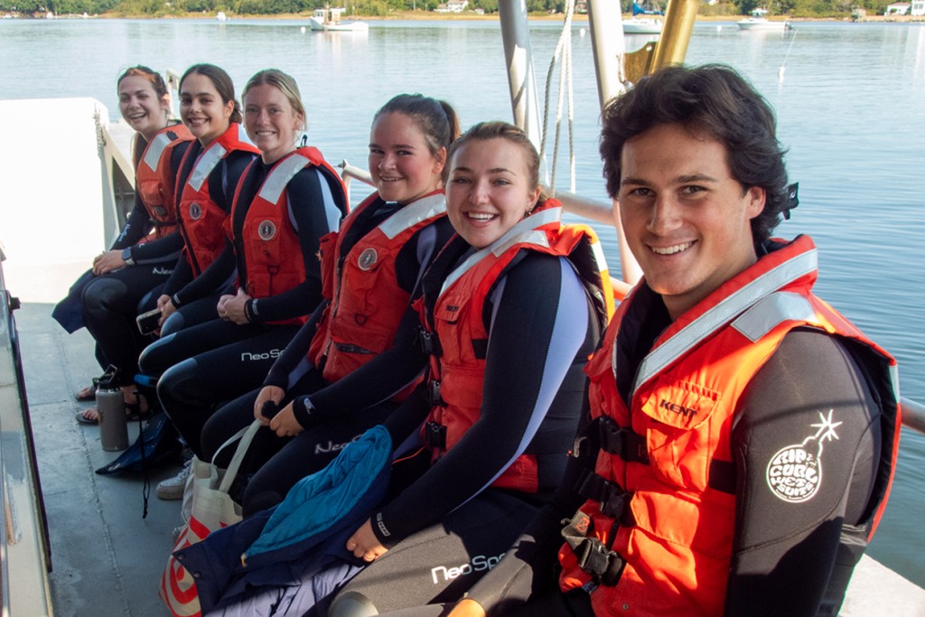 A group of students in orange life vests sit in a row on a U N E boat