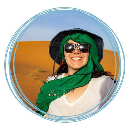 A student wearing a green scarf, sun hat, and sunglasses smiles while standing in a Moroccan desert