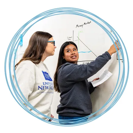 A student points to and discusses an economics graph on a white board with another student