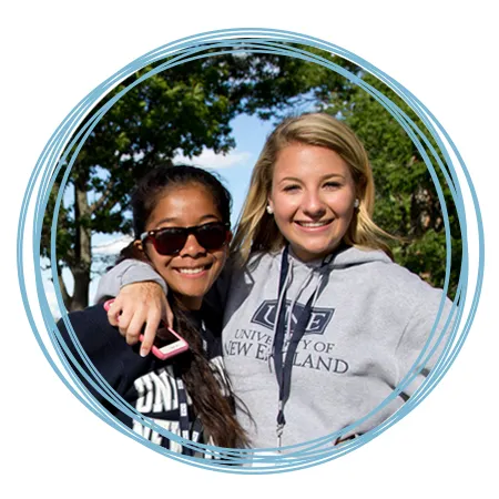 Two smiling students in U N E sweatshirts stand outdoors together on the Biddeford Campus