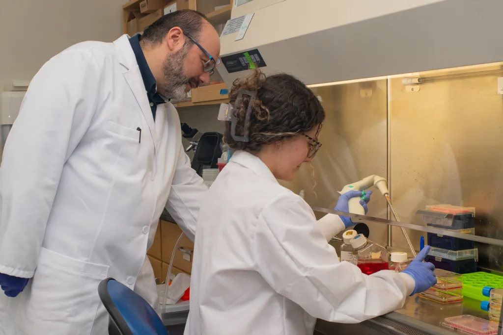 A professor watches over a student as they work with lab equipment