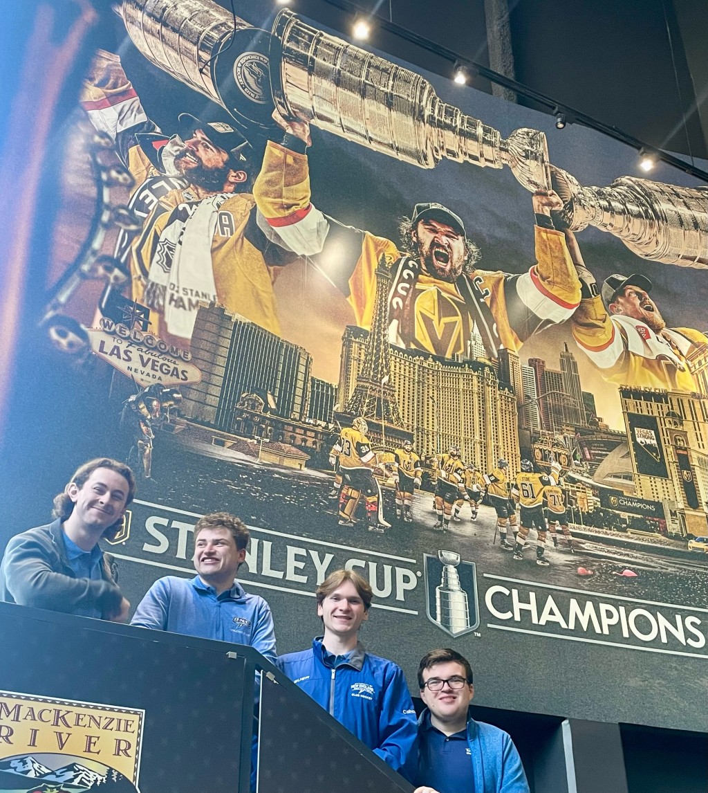Four male students pose in front of a large hockey advertisement