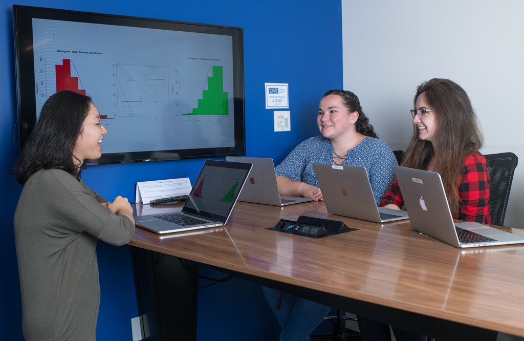 A group of data science students sit at a table with their laptops and graphs on a large monitor
