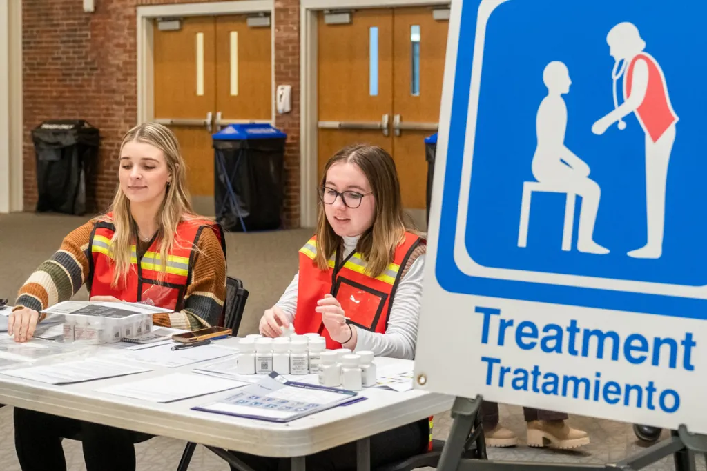Two nursing students sit with several forms at a table next to a sign that reads "Treatment"